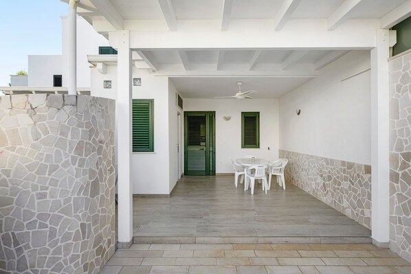 A covered outdoor area with a stone wall, a table with chairs, and a ceiling fan.
