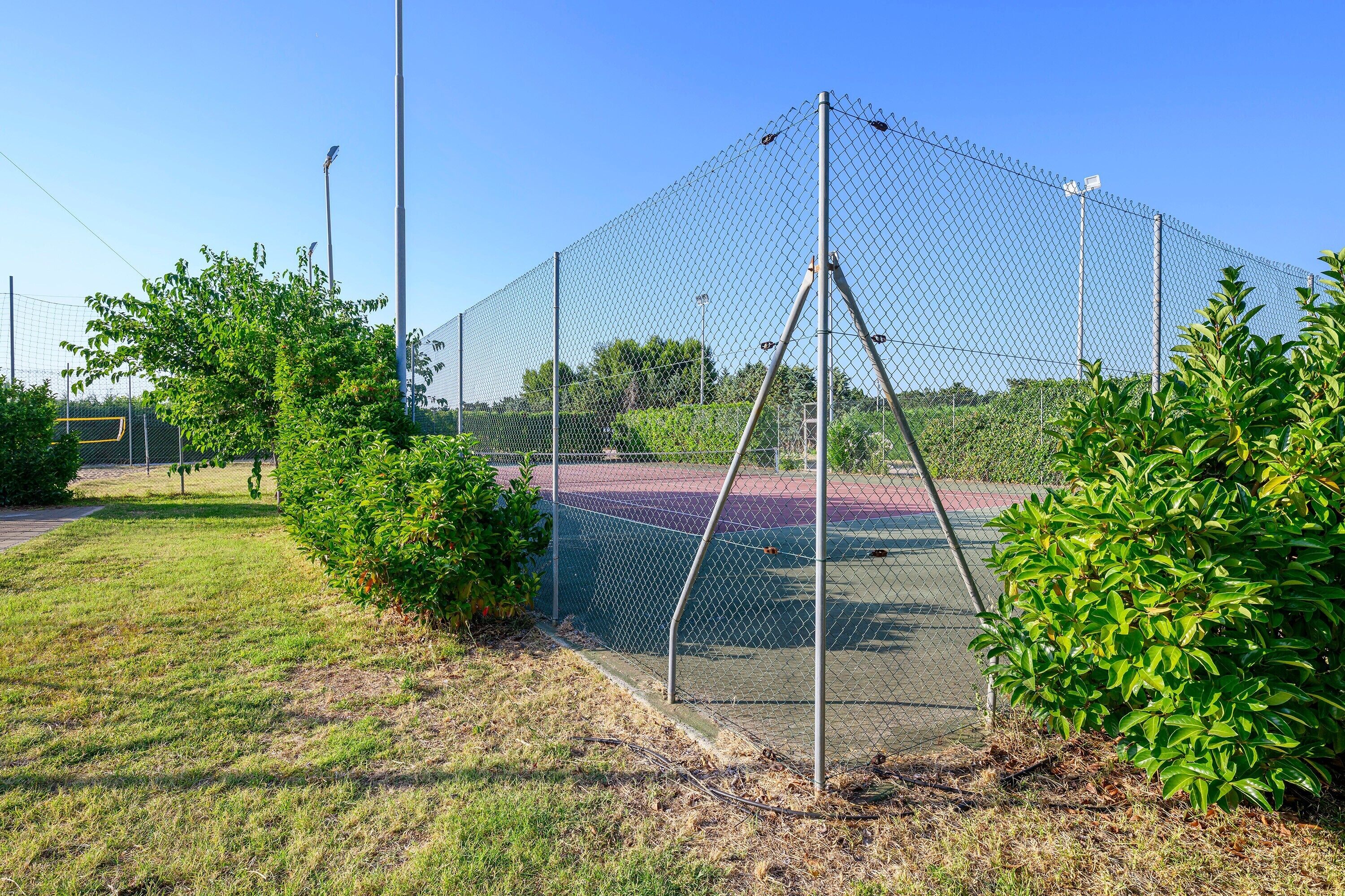 Ett stängsel av sammanlänkade metallkedjor omger en tennisplan, och ovanför syns en klar himmel.