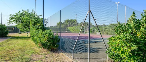 Ett stängsel av sammanlänkade metallkedjor omger en tennisplan, och ovanför syns en klar himmel.
