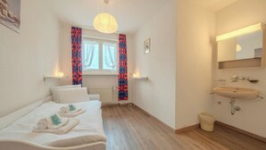 Habitación de hotel con cama, mesita de noche con toallas, ventana con cortinas, baño con lavamanos y espejo, y un radiador.