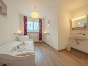 A hotel room with a bed, a bedside table with towels, a window with curtains, a bathroom with a sink and mirror, and a radiator. - Viktoria A22 by Arosa Vacations Appartement/Fewo, Dusche und Badewanne, 3 Schlaf by Interhome (Arosa)