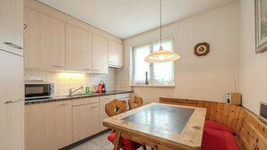 Cocina compacta con armarios de madera, un microondas y un lavabo. Hay una mesa de comedor con sillas y un banco con cojín rojo. Un reloj y una radio están en la pared.