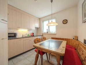 A compact kitchen with wooden cabinets, a microwave, and a sink. There's a dining table with chairs and a bench with a red cushion. A clock and a radio are on the wall. - Viktoria A22 by Arosa Vacations Appartement/Fewo, Dusche und Badewanne, 3 Schlaf by Interhome (Arosa)