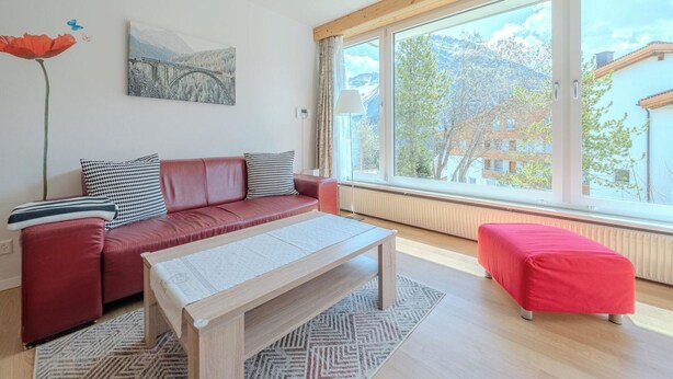 Una sala de estar moderna con un sofá rojo, una mesa de centro de madera y un ventanal que ofrece vistas a edificios y árboles.