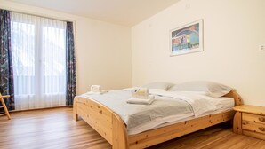 Habitación individual con cama de madera, mesita de noche de madera, ventana con cortinas traslúcidas y un cuadro en la pared.