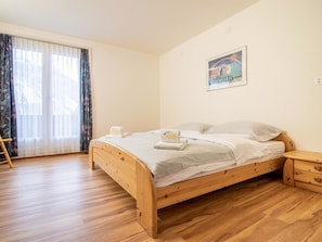 Ein Zimmer mit einem Einzelbett, einem hölzernen Nachttisch, einem Fenster mit leichtem Vorhang und einem Bild an der Wand.