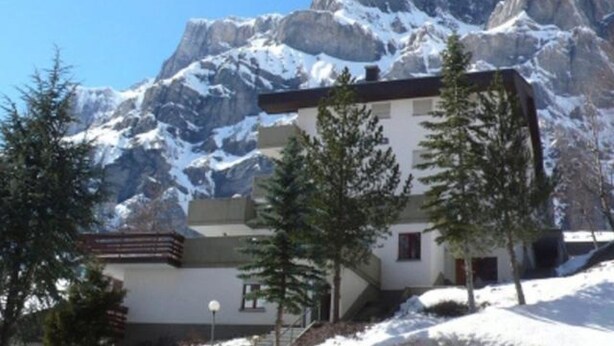 Un rifugio alpino con vista su cime innevate, un pontile di legno e una piccola scala che conduce all'ingresso.