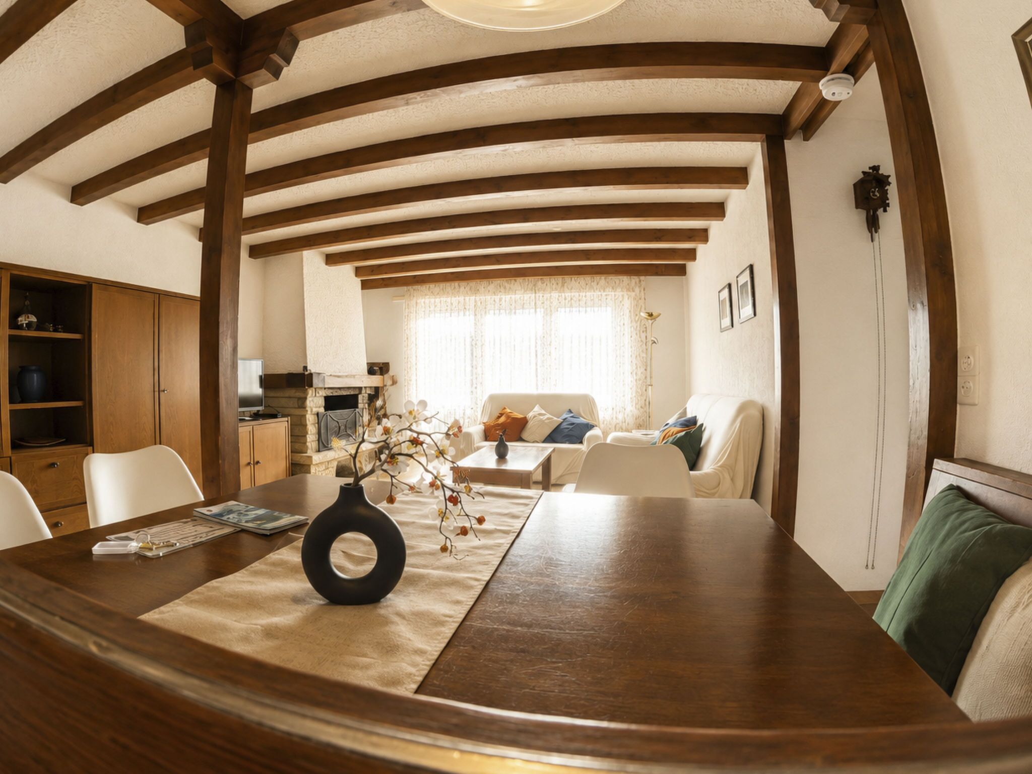 Un comedor con mesa de madera, sillas y un jarrón con flores. Se observa un sofá y una ventana con cortinas al fondo.