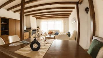 Un comedor con mesa de madera, sillas y un jarrón con flores. Se observa un sofá y una ventana con cortinas al fondo.
