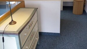 Une pièce comprenant un bureau, une lampe, une commode, une étagère et un petit tabouret.