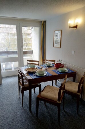 A dining area with a table set for four, chairs, a window with curtains, and a wall-mounted light fixture. - Fortuna 317 # 6-bed apartment, Dusche, WC 6-Bettwohnung, Dusche, WC by Interhome (Leukerbad)
