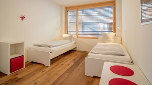 Une chambre moderne avec deux lits simples, un plancher en bois, une fenêtre avec des stores et une petite décoration rouge sur le mur.