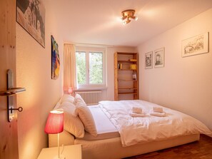 A bedroom with a bed, a bedside table, a lamp, a radiator, a window with curtains, a shelf with books, and framed pictures on the wall. - Latschüel 3.OG by Arosa Vacations Appartement/Fewo, Dusche und Bad, WC, 2 Schlaf by Interhome (Arosa)