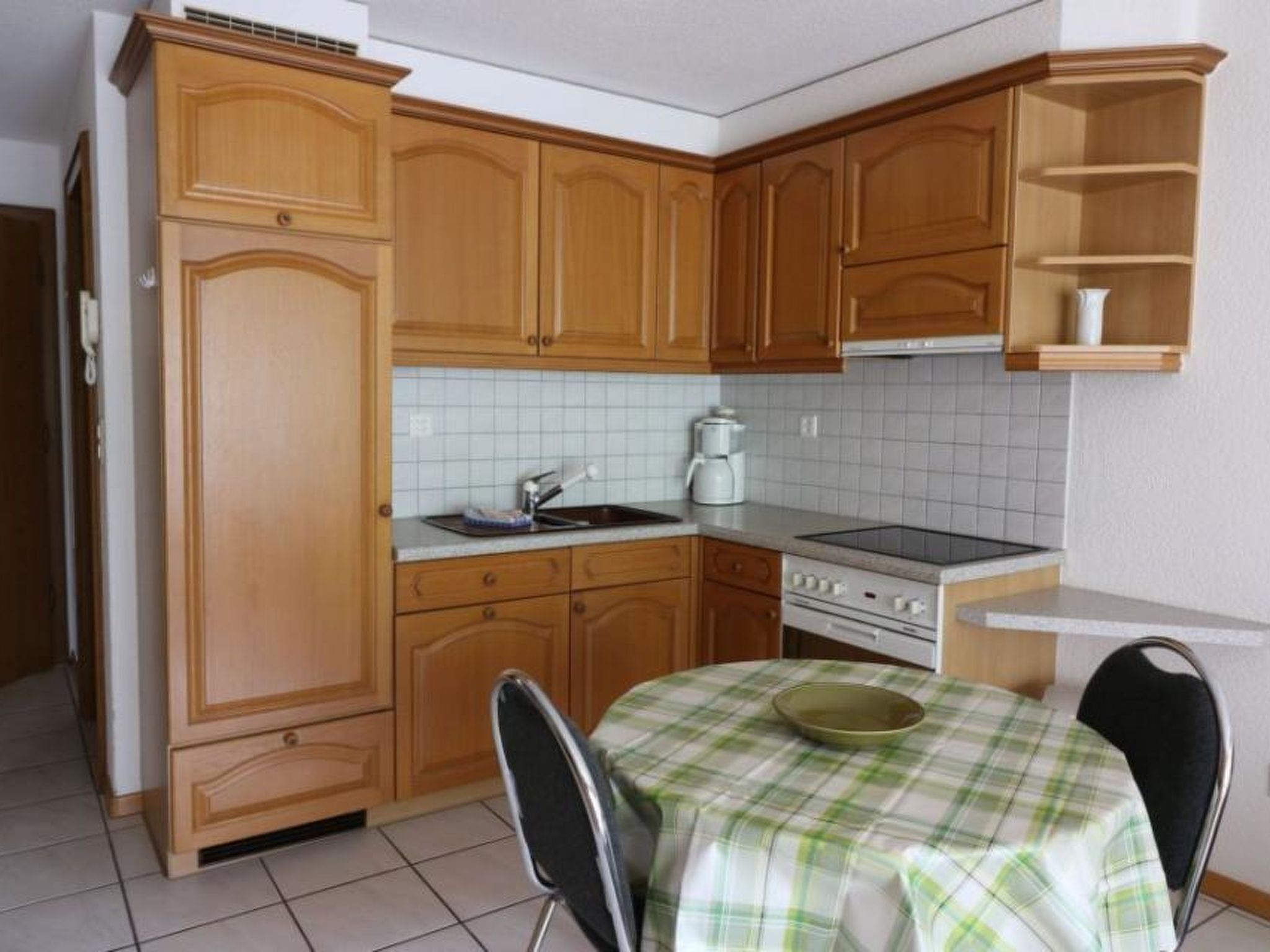 A compact kitchen with wooden cabinets, a white tiled backsplash, a stove, and a small dining table with a checkered tablecloth.