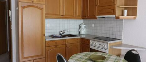 A compact kitchen with wooden cabinets, a white tiled backsplash, a stove, and a small dining table with a checkered tablecloth.