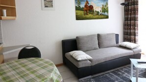 Olohuone, jossa on sohva, ruudullisella pöytäliinalla katettu pöytä, hevosten kuva ja ikkuna, jossa on verhot.