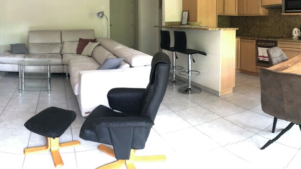 A living room with a sofa, a black chair, a small table, and a kitchen area with cabinets and a countertop. - Edelweiss D 02 # 4-bed apartment, Dusche, WC 4-Bettwohnung, Dusche, WC by Interhome (Leukerbad)