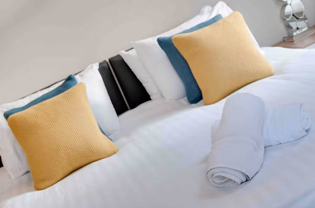 A neatly made bed with white bedding, a rolled towel, and pillows with blue and yellow stripes.