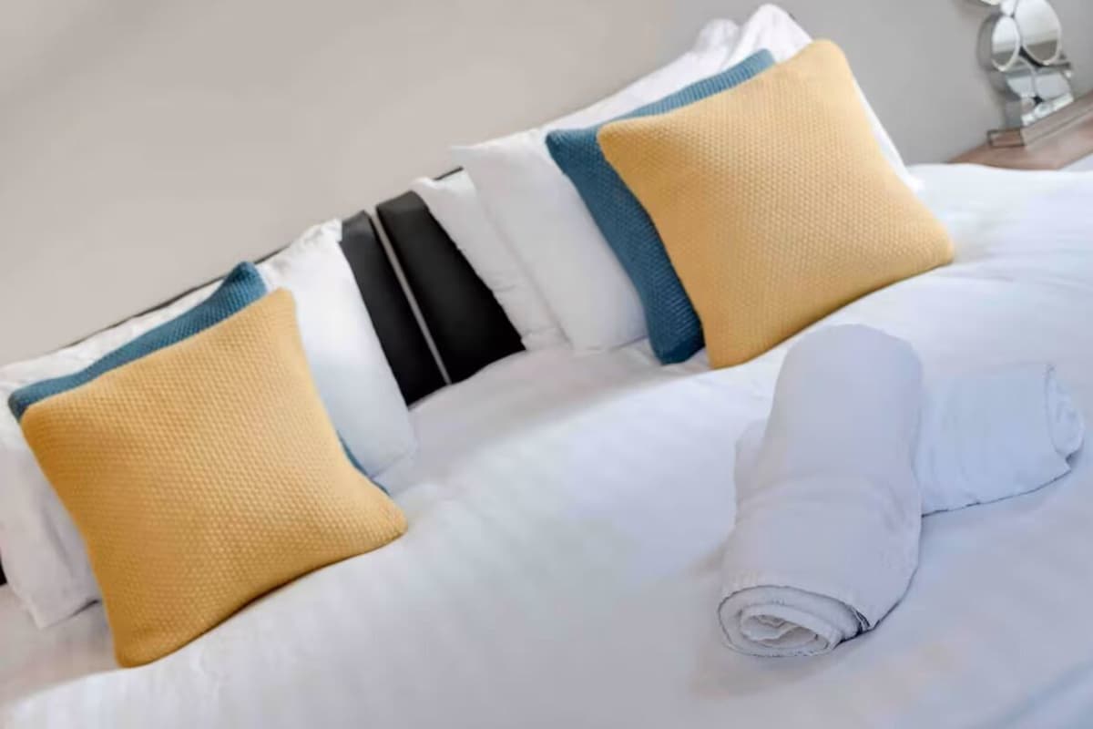 Un letto rifatto con cura, lenzuola bianche, un asciugamano arrotolato e cuscini a righe blu e gialle.
