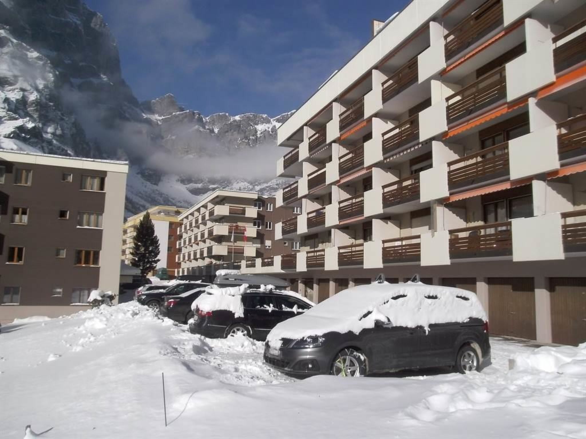 A multi-story building with balconies, parked cars covered in snow, and mountains in the background.