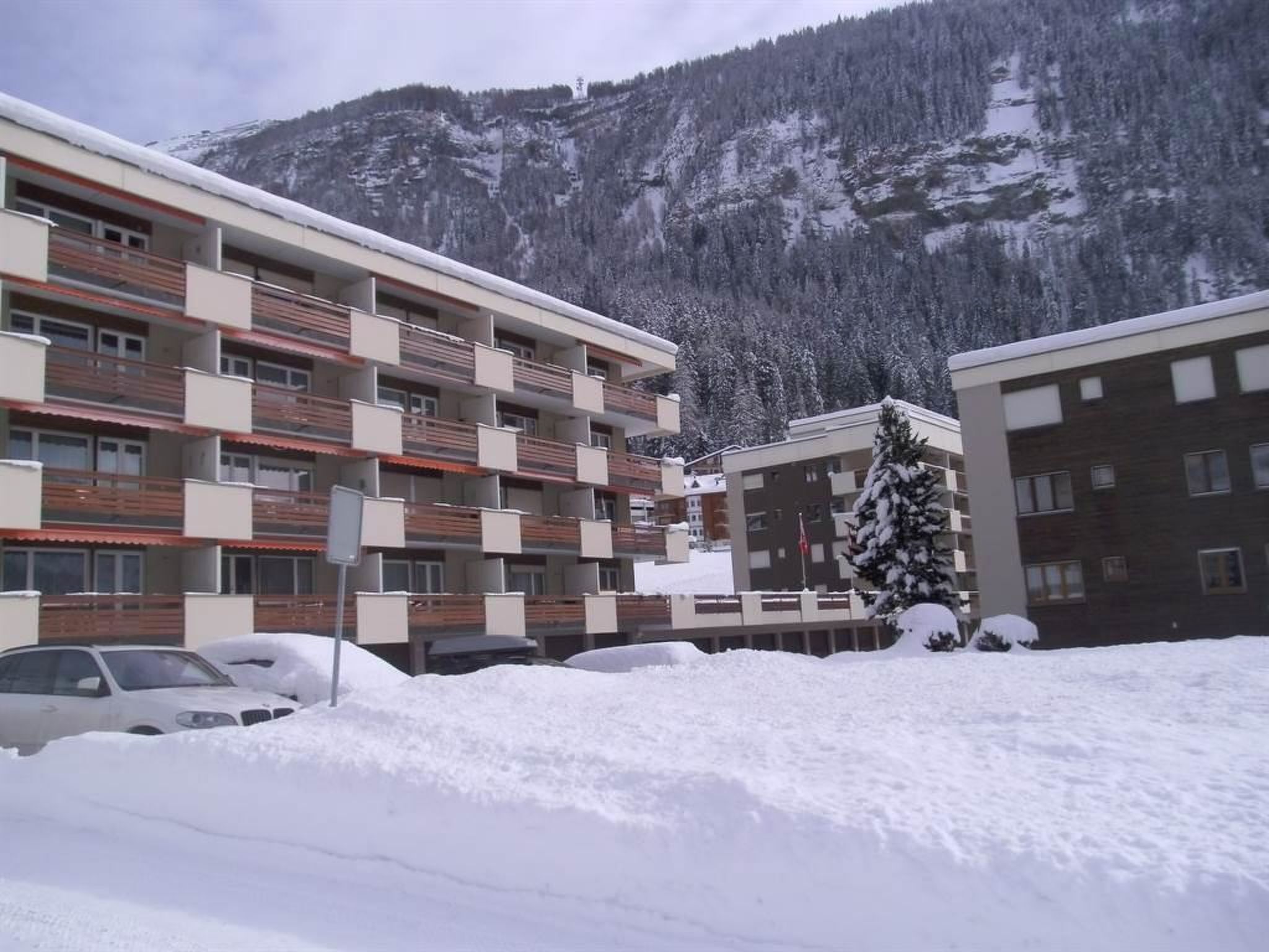 Et hotel med flere etager og balkoner, med et snedækket bjerg i baggrunden og en parkeret bil.