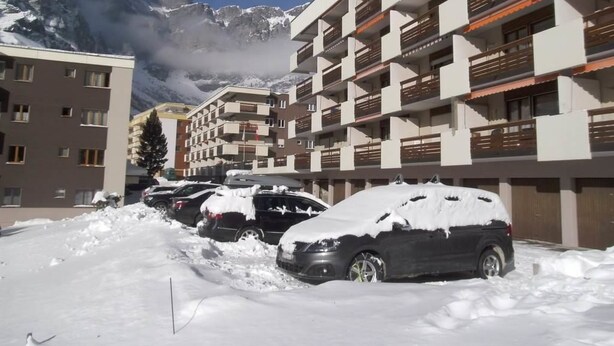 Un edificio de varios pisos con balcones, coches aparcados cubiertos de nieve y montañas al fondo.