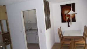Una habitación con una mesa de comedor, sillas, una lámpara colgante, un cuadro y una cocina visible a través de una puerta abierta.