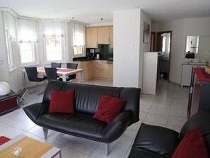 A modern living room with a black leather sofa, a dining area with a table and chairs, and a kitchen with wooden cabinets and appliances. - Edelweiss C 31 # 4-bed apartment, Dusche, WC 4-Bettwohnung, Dusche, WC by Interhome (Leukerbad)