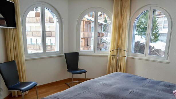 Hotellihuone, jossa on sänky, kaksi tuolia ja suuret ikkunat, joissa on verhot.
