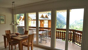Un comedor con mesa y sillas, ventanas amplias con vista a edificios y montañas, y un balcón con barandilla.