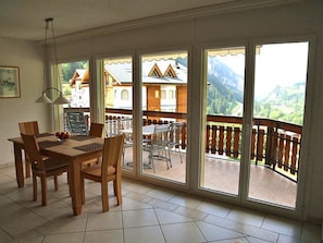 Ein Essbereich mit Tisch und Stühlen, große Fenster mit Blick auf Gebäude und Berge sowie ein Balkon mit Geländer.