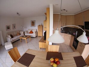 Ein modernes Wohnzimmer mit Sofa, Couchtisch und Essbereich mit Tisch und Stühlen. Es gibt eine Küchenzeile mit Schränken und Herd.
