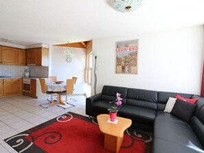 Ein Wohnzimmer mit einer schwarzen Couch, einem kleinen Holztisch und einem roten Teppich. Im Hintergrund ist eine Küche mit Holzschränken und Fliesenboden zu sehen.