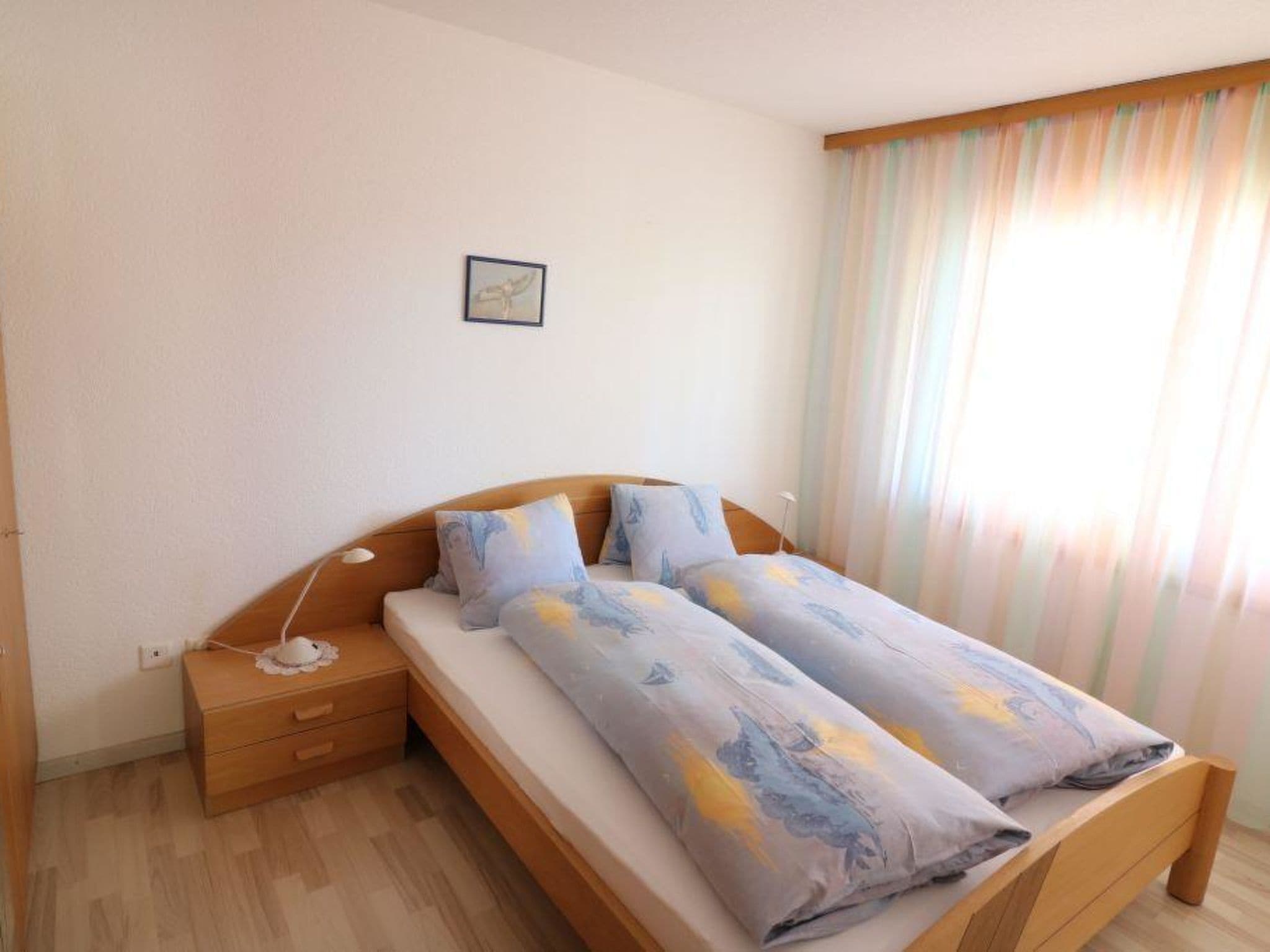 A single bed with a wooden frame and a headboard, a bedside table with a lamp, and a window with sheer curtains.