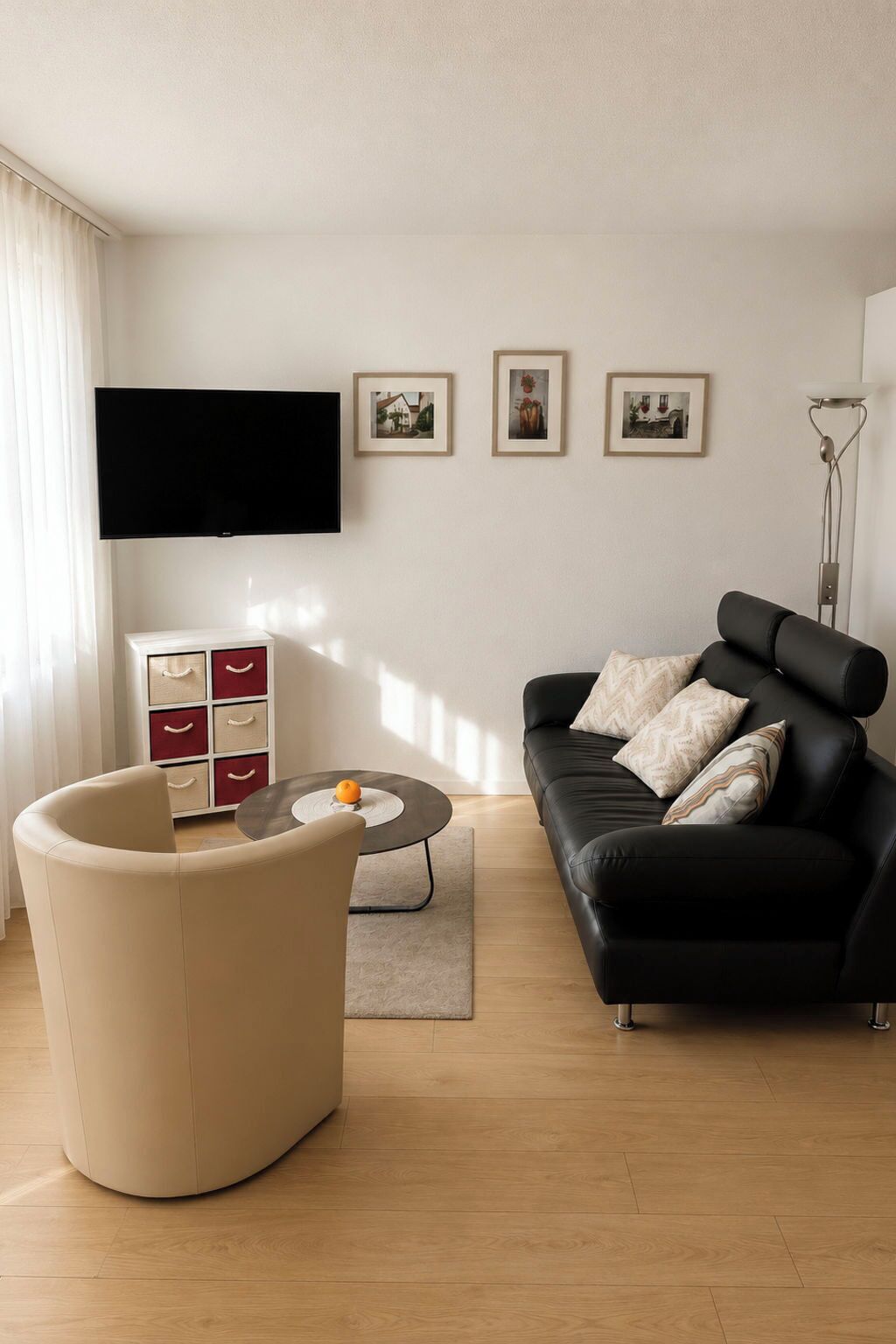 Ein modernes Wohnzimmer mit einer schwarzen Couch, einem runden Couchtisch, einer weißen Kommode mit roten Schubladen und einem flachbildfernseher an der Wand.
