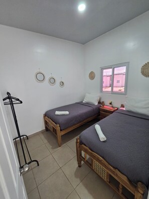 Ein Zimmer mit zwei Holzbett, ein Fenster mit Ausblick, einem an der Wand befestigten Wäscheständer und einer Deckenleuchte.