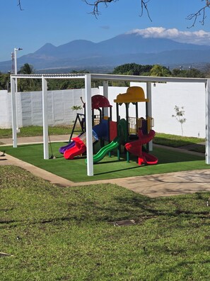 Ein Spielplatz mit Rutschen und Klettergerüsten, umgeben von einem weißen Zaun und Gras, mit Bergen im Hintergrund.