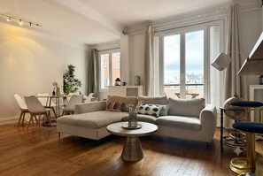 A living room with a sofa, round coffee table, dining table, chairs, and a view through large windows. - The Pastor's White Cocoon - Apartment with a View of the Eiffel Tower C08 (Paris)