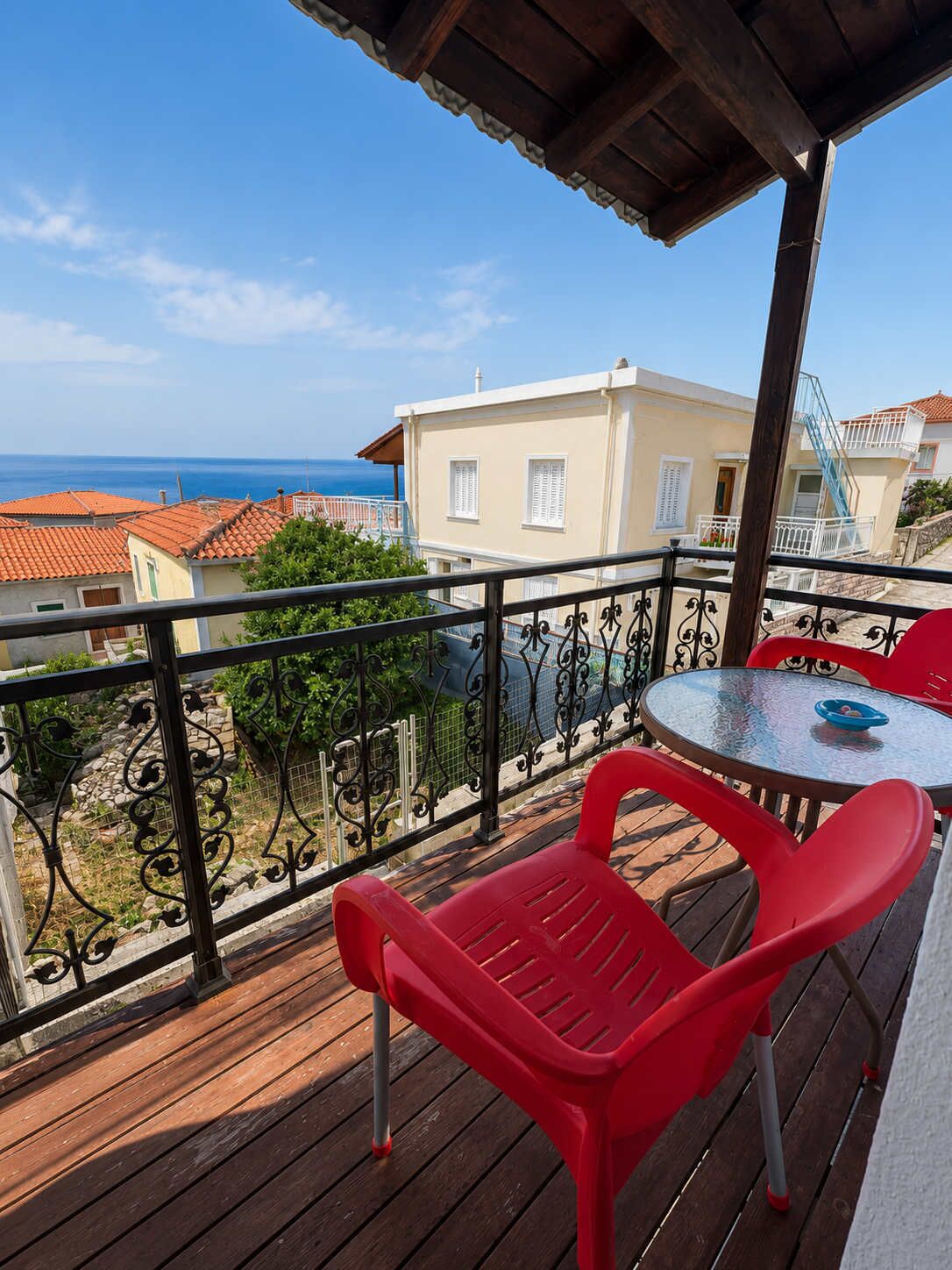A balcony with a table and chairs, overlooking a coastal town with terracotta roofs.