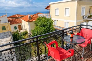 A balcony with red chairs and a table, overlooking a coastal town with terracotta roofs and a clear sky. - Little nest - Serene and Cozy Samos Retreat (Koumeika Samou)