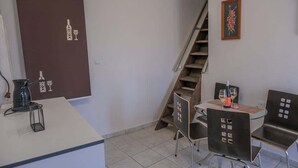 Una habitación pequeña con kitchenette, un comedor con dos sillas, una escalera y un cuadro enmarcado en la pared.