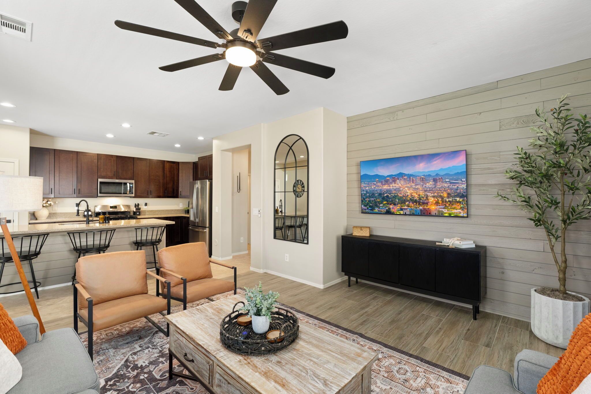 A modern living room with a dining area, a kitchen, a ceiling fan, a mounted TV, and a potted plant.