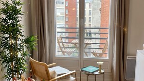 Ein Zimmer mit Holzboden, einem runden Tisch mit einer Vase roter Beeren, einem Stuhl, einem kleinen Tisch mit einem Buch und Blick auf Gebäude durch große Fenster.