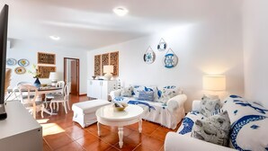 Ein Wohnzimmer mit weißem Sofa, einem runden Couchtisch, einem Essbereich mit Stühlen und Tisch sowie einer Küche mit Schränken.
