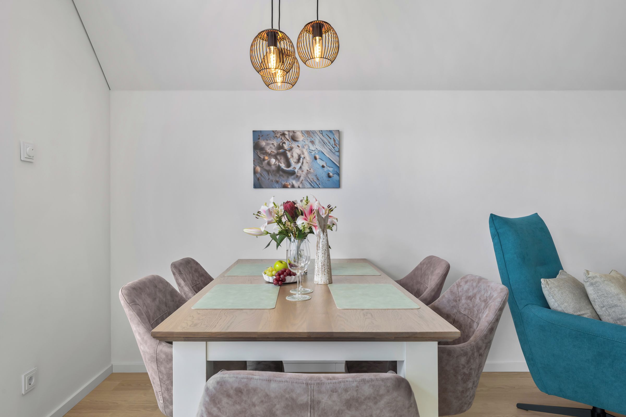 A dining area with a wooden table, cushioned chairs, a vase with flowers, and a painting on the wall.