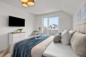 A modern bedroom with a flat-screen TV, a bedside table with a plant, a window with a view of houses, and a bed with a blue blanket. - Perfekte Welle - balcony, electric car charging station, private parking (Kappeln)