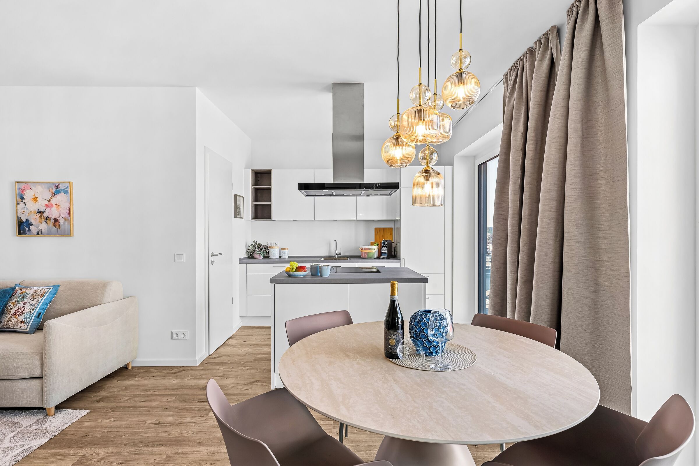 A modern kitchen with white cabinets, a dining area with a round table, and a living room with a sofa.