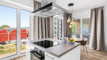 Et moderne køkken med en central ø, indbygget ovn og et stort vindue med udsigt til et rødt ladebygning og grøn bevoksning.