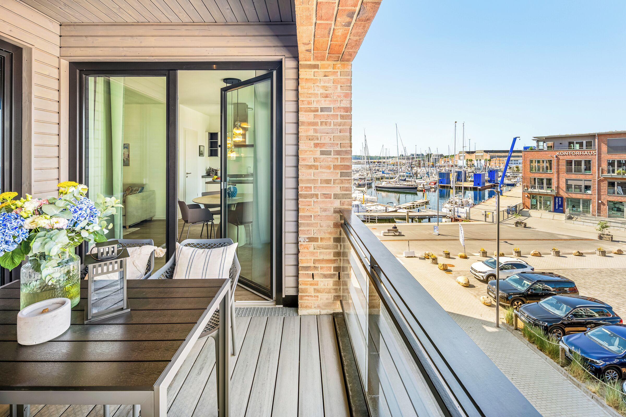 Ein Balkon mit Holzboden, Tisch und Stühlen mit Blick auf einen Yachthafen, in dem sich Boote und ein Gebäude mit der Aufschrift 'KONTORHAUS' befinden.