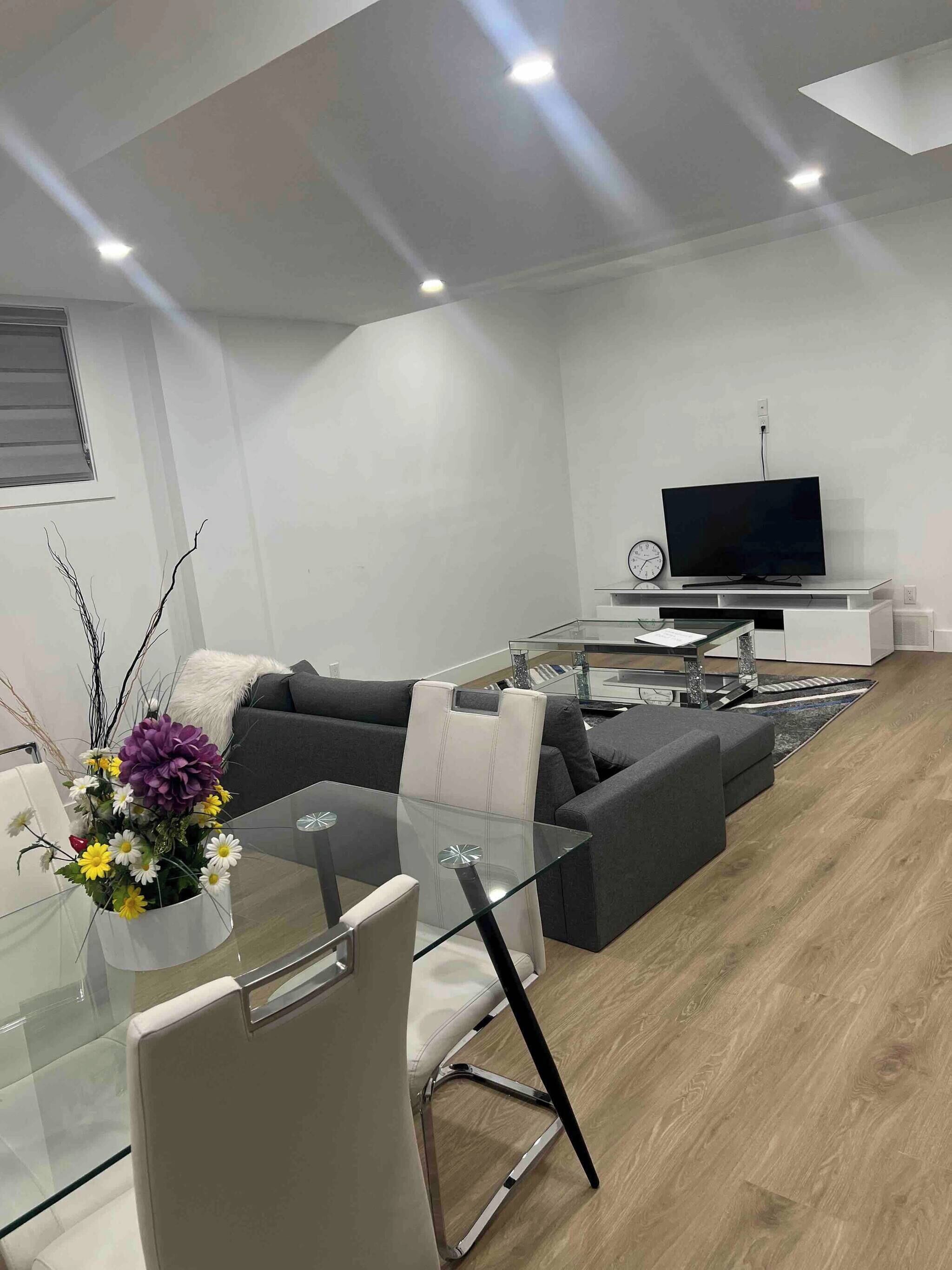 Een moderne woonkamer met een glazen eettafel, een bank, een televisie en een decoratieve vaas met bloemen.